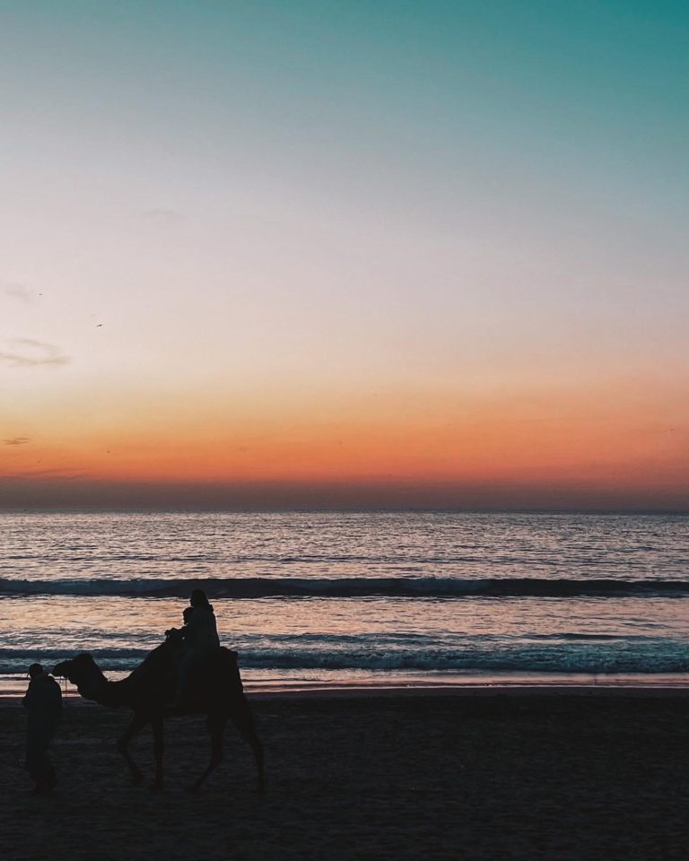 the sunset in Agadir showing blue, white, orange and yellow tones fading behind the sea front with a silhouette of someone riding a camel in the bottom left