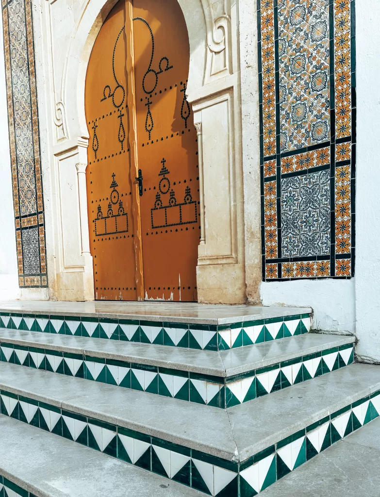 A yellow/mustard coloured Tunisian door in Sidi Bou Said with black geometric patterns. Further abstract style artwork are on the walls next to the door while geometric patterns are on the short staircase leading to the door