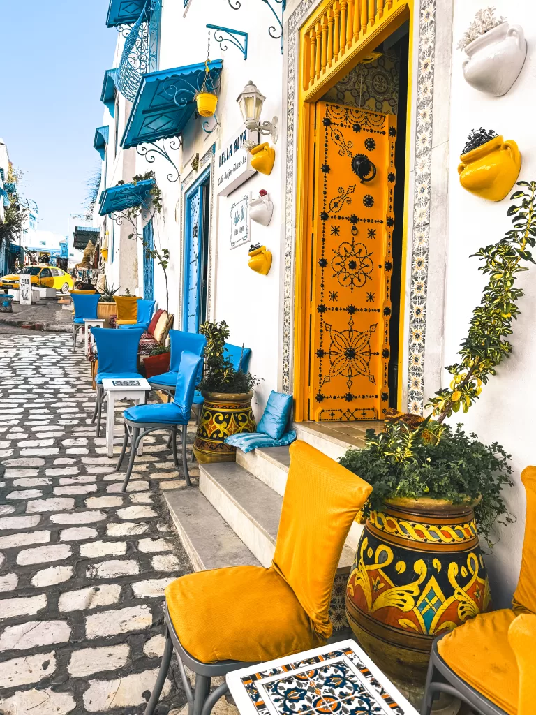 A colourful street featuring blue and yellow Tunisian doors, architecture and geometric patterns with yellow and boot seating outside of a cafe