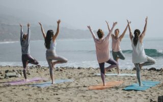 Five women practicing yoga on a sandy beach with ocean waves in the background.