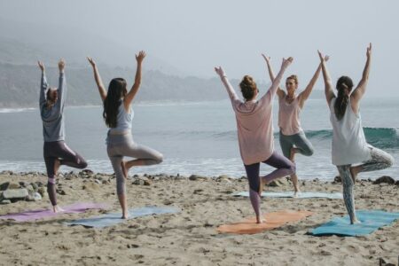 Five women practicing yoga on a sandy beach with ocean waves in the background.