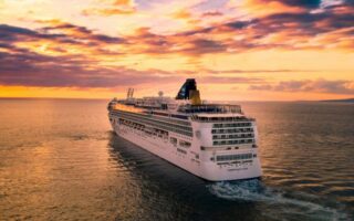 A large cruise ship sailing in calm waters during sunset, with orange and purple skies.