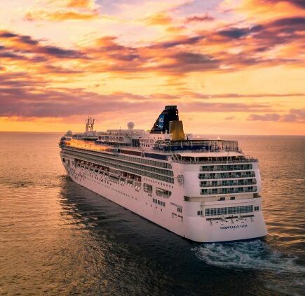 A large cruise ship sailing in calm waters during sunset, with orange and purple skies.