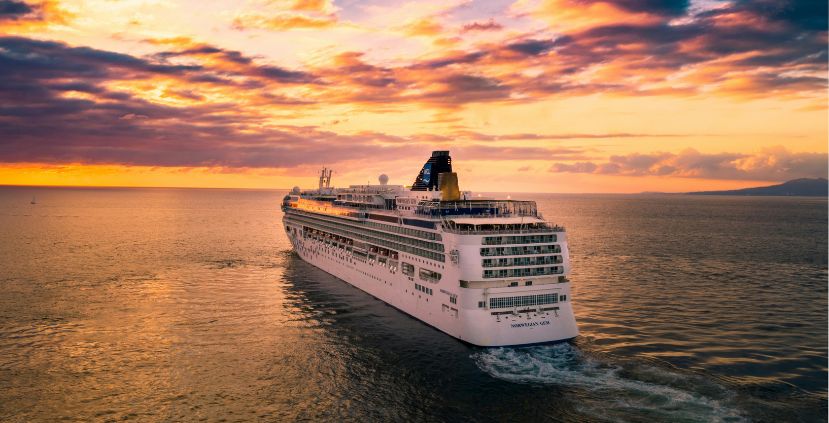 A large cruise ship sailing in calm waters during sunset, with orange and purple skies.